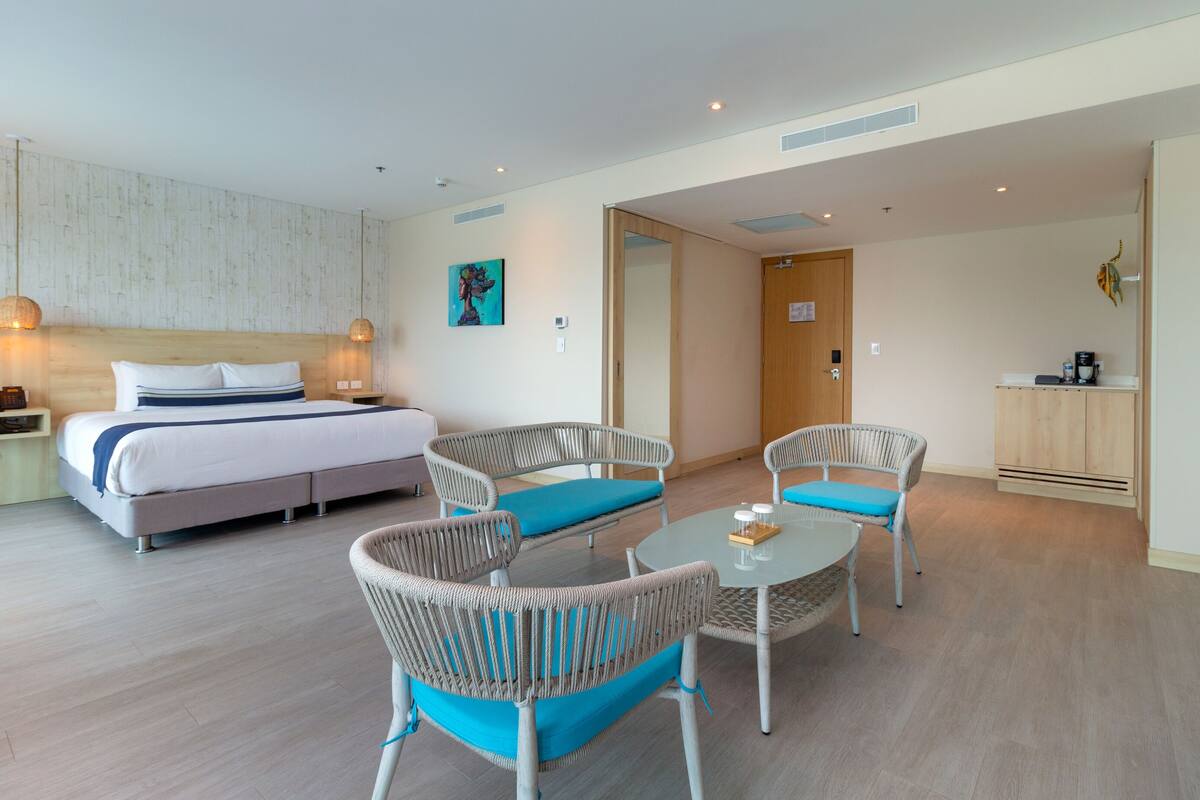 Une chambre d’hôtel avec un lit, deux fauteuils en osier dotés de coussins bleus, une petite table ronde, un meuble en bois et une porte donnant sur une autre pièce.