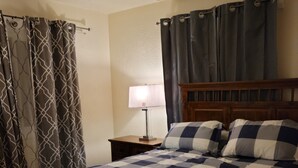 Un dormitorio con cabecera de cama de madera, colcha a cuadros, mesita de noche con lámpara y cortinas.