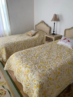 Una habitación con dos camas, cada una con una colcha estampada, una mesita de noche con lámpara y una ventana con cortinas.