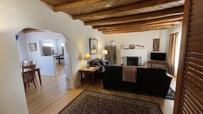 Una sala de estar con un sofá negro, piso de madera y televisión. Hay una zona de comedor con sillas y una mesa.