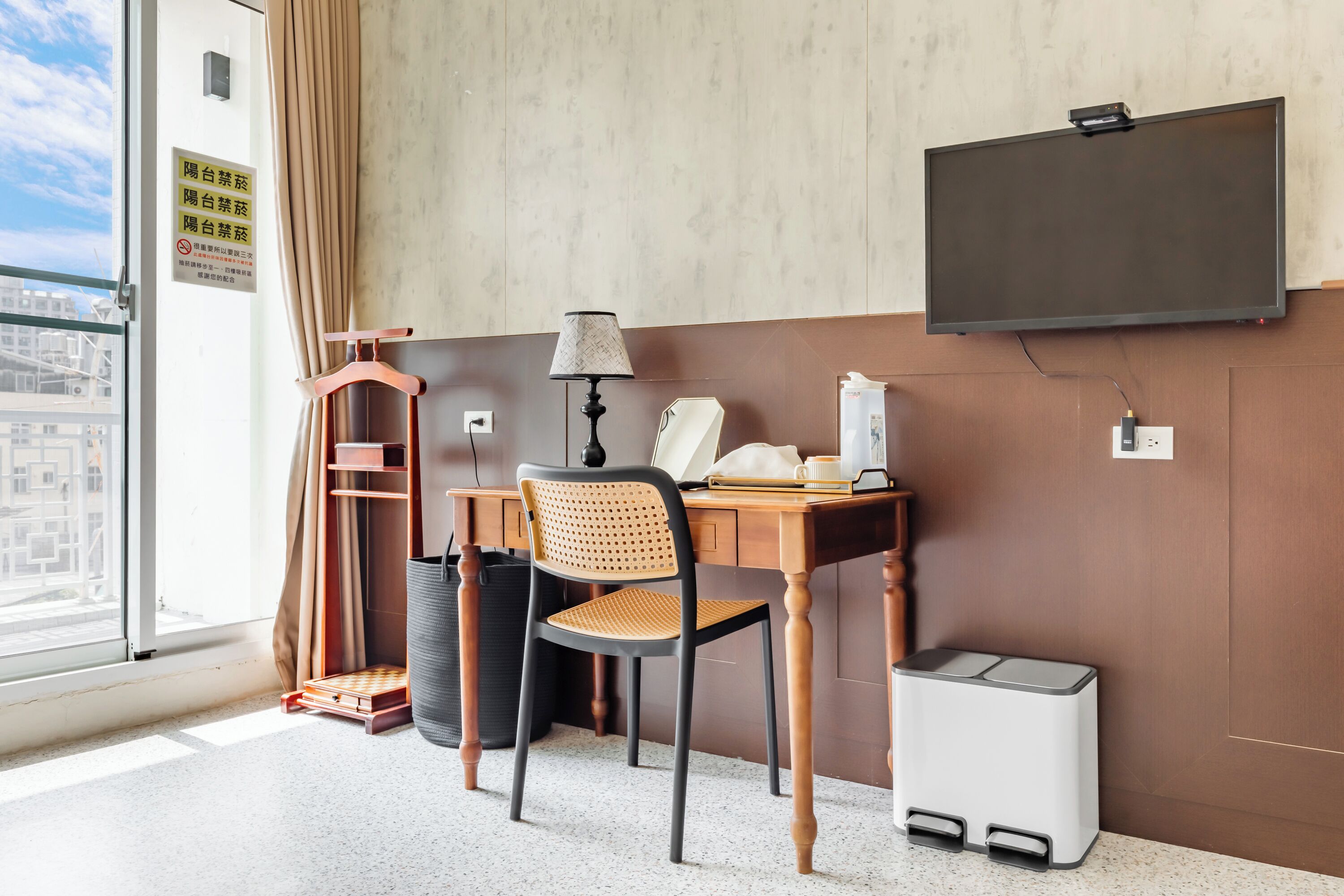 Habitación de hotel con un escritorio de madera, una silla, una lámpara, un televisor montado en la pared, una ventana con cortinas y vistas a la ciudad.
