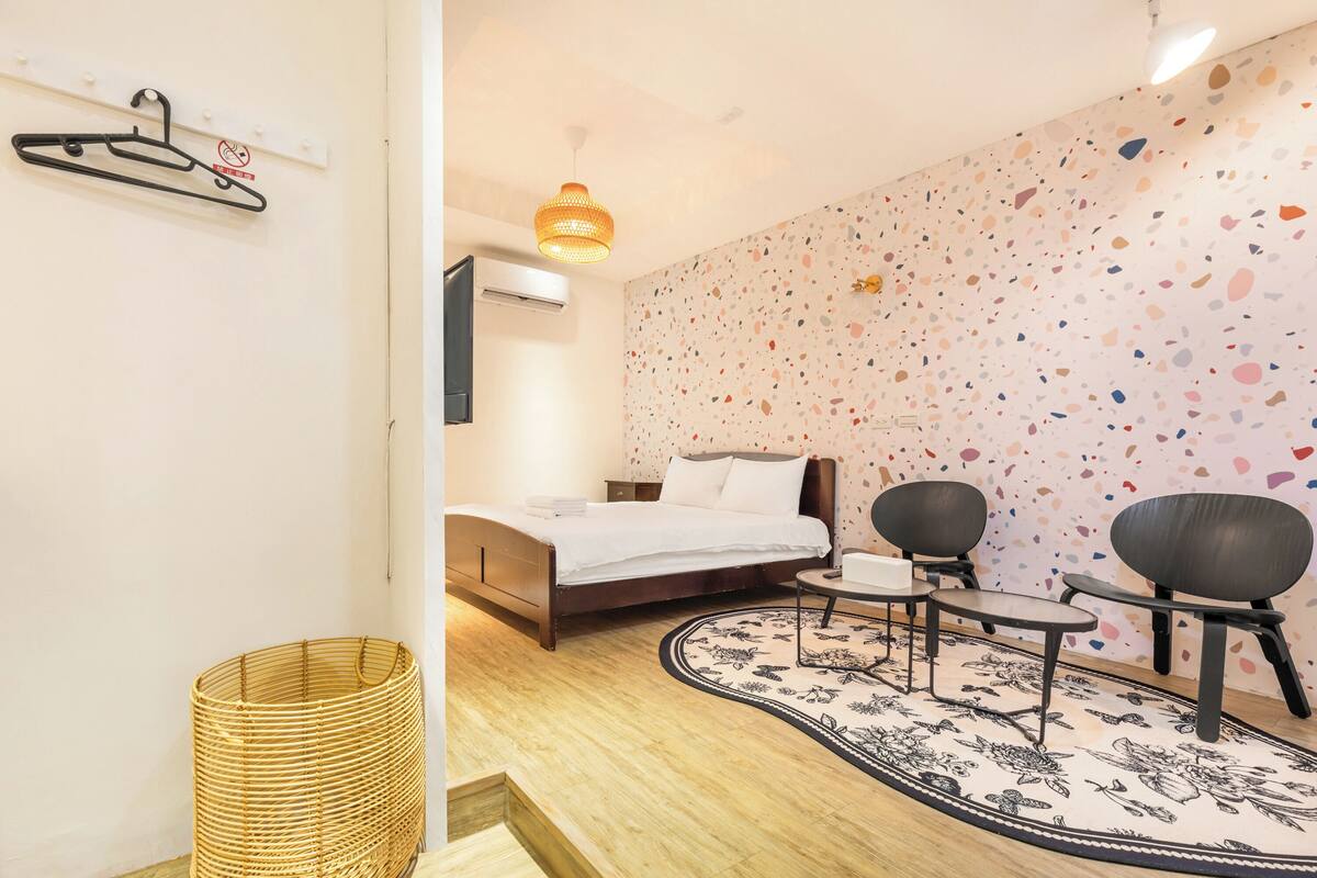 A bedroom with a bed, a bedside table, two chairs, a small table, a hanging light fixture, and a patterned wall.