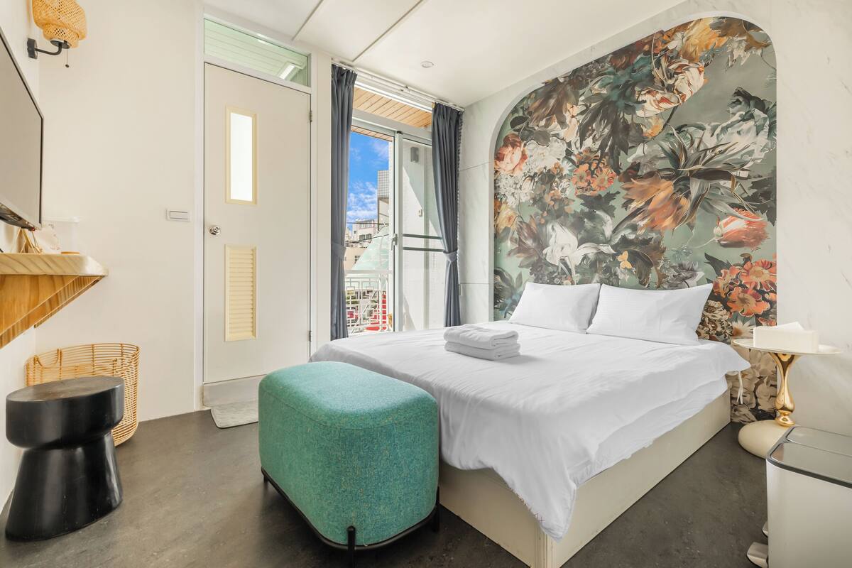 A hotel room with a bed, a window with curtains, a wall with a floral mural, a small table, and a chair.