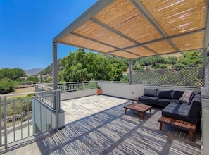 Relax on the spacious outdoor terrace with a pergola, sofa, and bench, surrounded by lush greenery and a distant view.