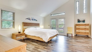 Een slaapkamer met een houten bed, nachtkastje, dressoir en een stoel. Er zijn twee ramen en een deur met uitzicht op groen.