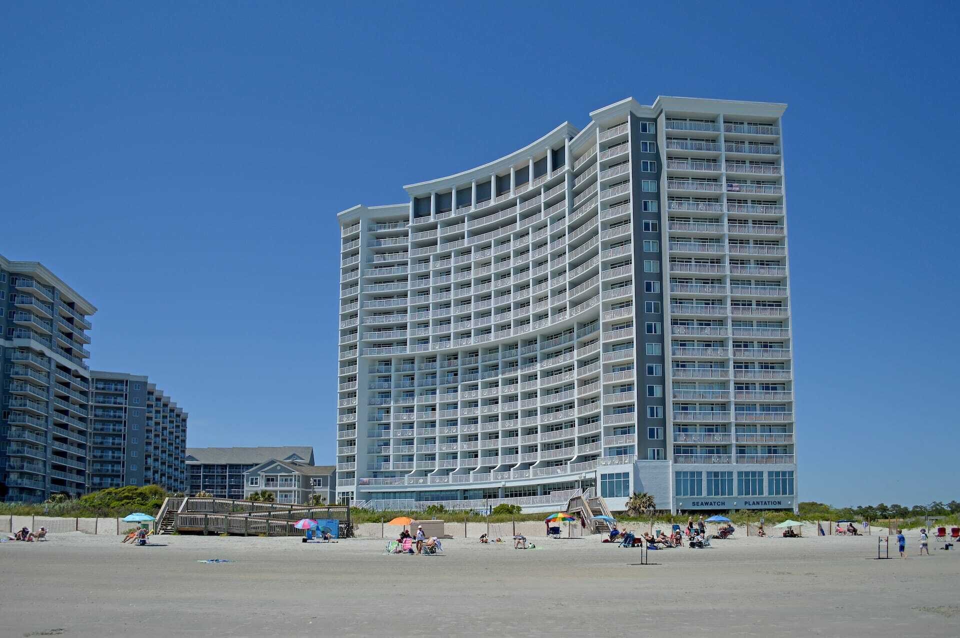 Un grand hôtel avec de nombreux balcons, situé juste à côté d’une plage où se trouvent des gens et des parasols.