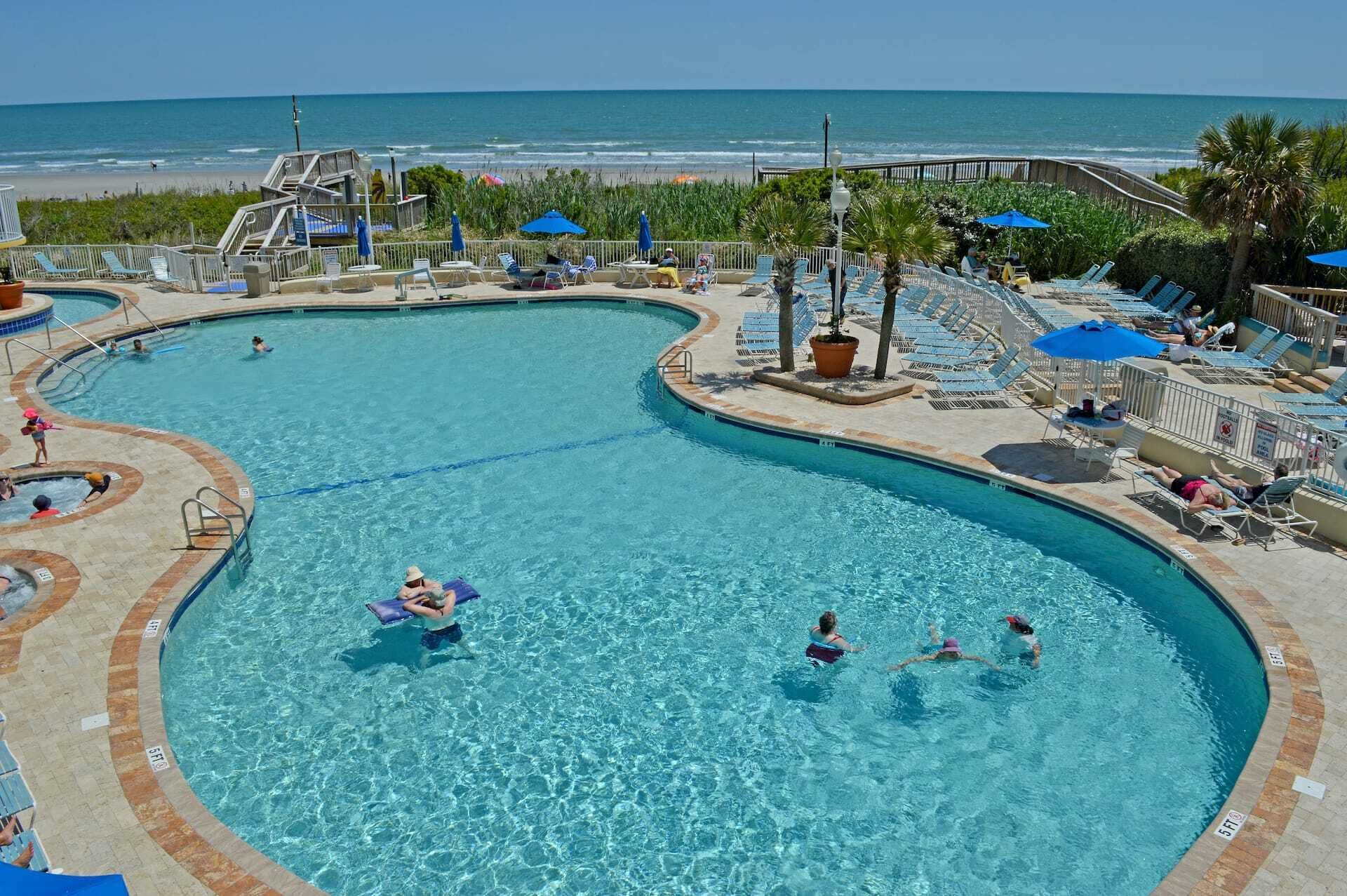 Une grande piscine à eau claire et bleue, de forme incurvée, entourée de chaises longues et de parasols.