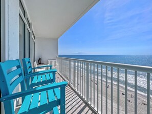 Un balcon avec des chaises turquoise et une balustrade blanche donnant sur une plage où se trouvent des gens et une statue.