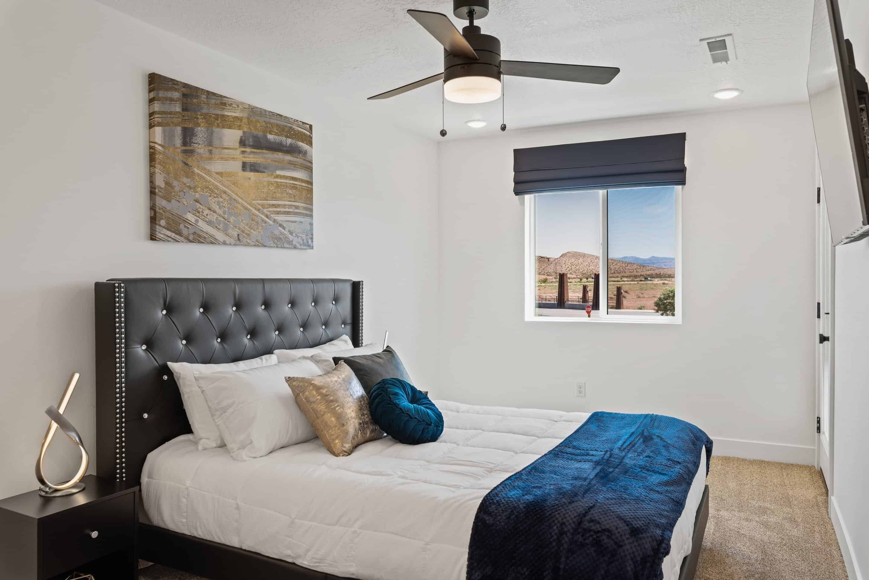 Une chambre à coucher avec une tête de lit capitonnée, un lit recouvert d’une literie blanche et bleue, une fenêtre donnant sur un paysage désertique et un ventilateur de plafond.