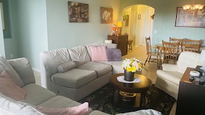 Een woonkamer met een bank, fauteuils, een salontafel met bloemen, een eethoek met stoelen en een kroonluchter.