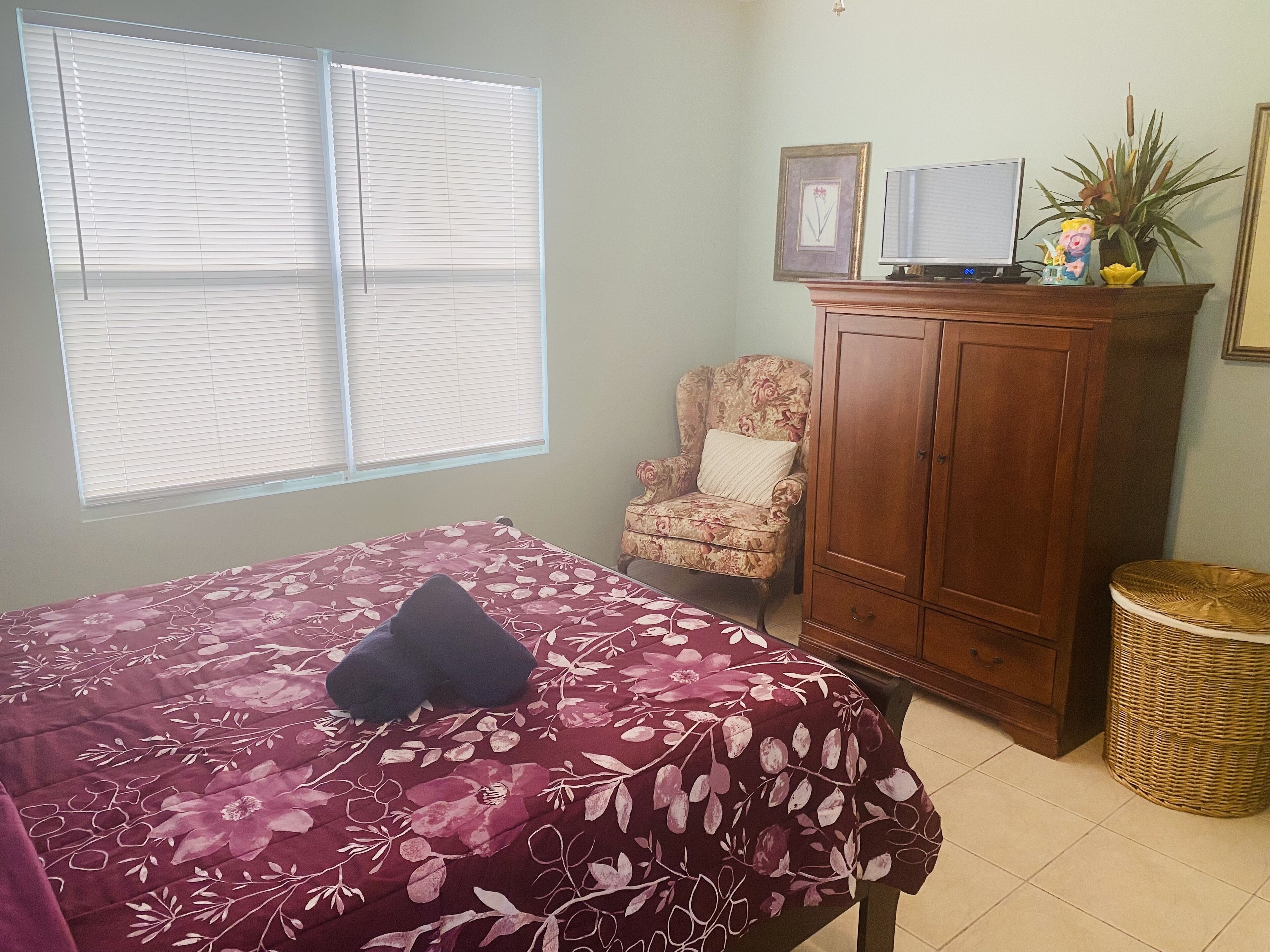 Un dormitorio con una cama, una silla, un armario de madera, un televisor y una ventana con persianas.