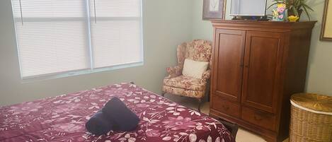 Un dormitorio con una cama, una silla, un armario de madera, un televisor y una ventana con persianas.