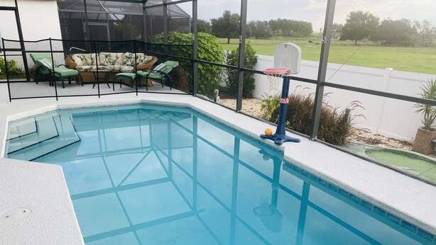 Een zwembad met een basketbalring, omringd door een overdekt terras met stoelen en een bank.