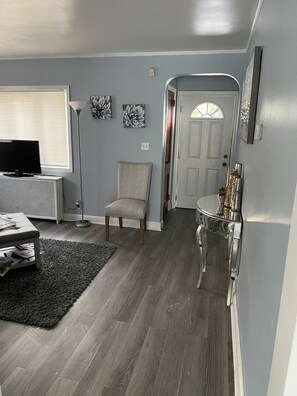 Un salón con un sofá gris, un televisor, una lámpara de pie y una alfombra. Hay una puerta que da a otra habitación.