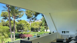 Un área de comedor al aire libre con una mesa dispuesta para dos personas, rodeada de sillas, un banco acolchado y vistas a un jardín con árboles y una montaña.