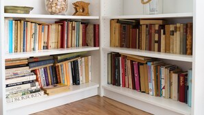 Una estantería con varios libros, un tarro de cristal con conchas y una escultura de madera.