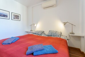 Een netjes opgemaakt bed met een rode sprei, twee kussens en twee gevouwen handdoeken. Twee wandlampen, een witte airconditioner en twee ingelijste foto’s hangen aan de muur.