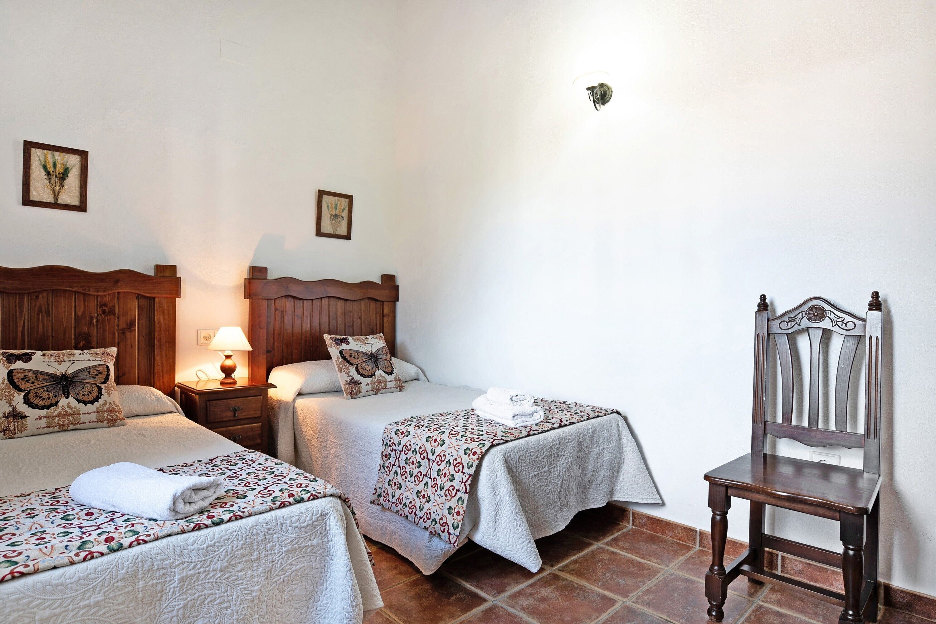 Una camera da letto con due letti, una sedia di legno, un comodino con una lampada e un punto luce a parete.