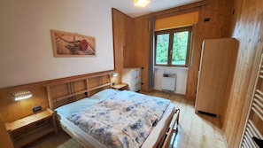 A wooden room with a bed, a dresser, a radiator, and a window with a view of greenery. - Apartment Oleandro Casa Demonti with Mountain View, Pool, Terrace & Wi-Fi (Tignale)