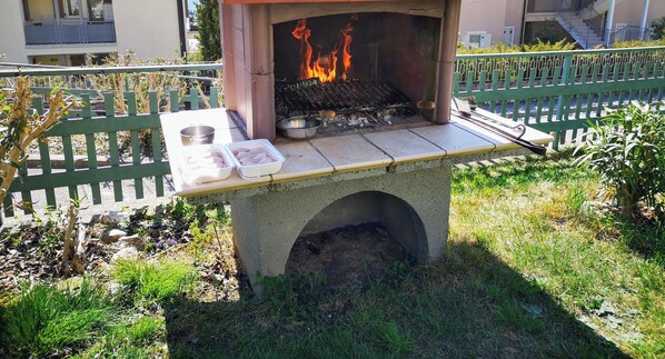 An outdoor grill with flames, a tray with food, and a bench. - Apartment Oleandro Casa Demonti with Mountain View, Pool, Terrace & Wi-Fi (Tignale)