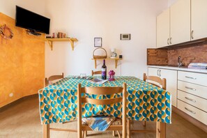 Un coin repas avec une table recouverte d’une nappe à motifs, des chaises en bois, un téléviseur fixé au mur et un espace cuisine comprenant des placards et un évier.