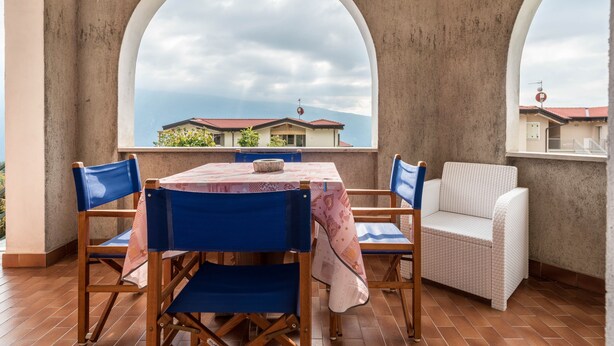 Eine Terrasse mit Tisch und Stühlen, ein weißes Korbsitzsofa und Blick auf ein Haus durch ein großes, rundes Fenster.