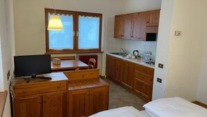 A compact room with a wooden desk, a bed, a kitchenette, and a window with curtains. - Monterosa Residence Hotel (Macugnaga)