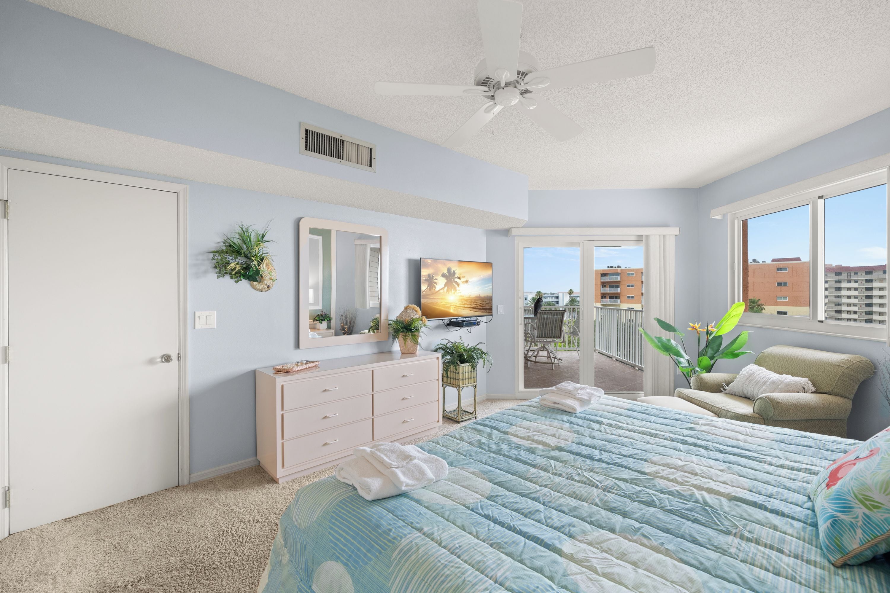 Une chambre avec un grand lit, un ventilateur de plafond, une commode, une télévision et un balcon agrémenté de plantes.