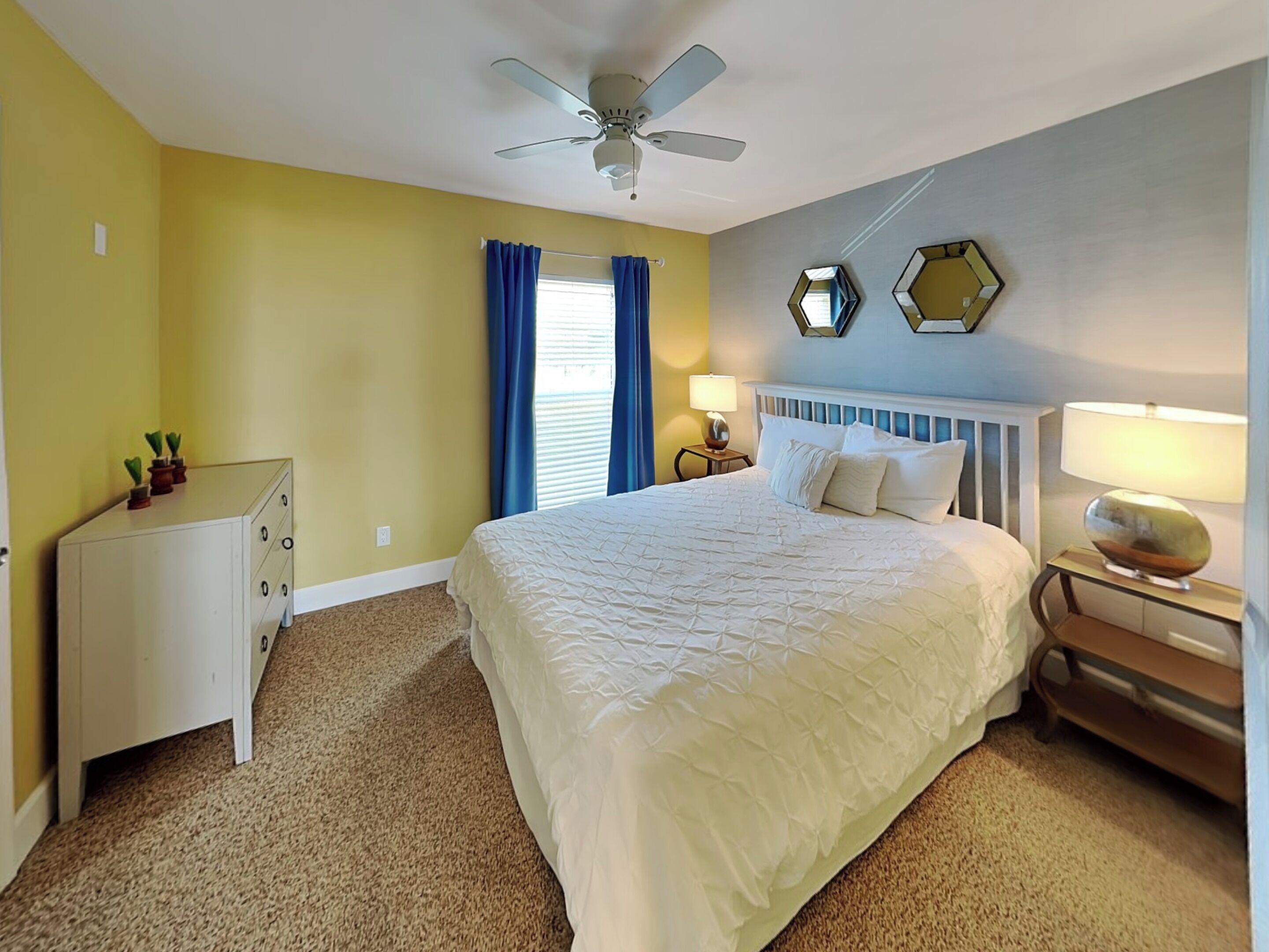 A bedroom with a bed, a dresser, a ceiling fan, a nightstand with a lamp, and a window with curtains.