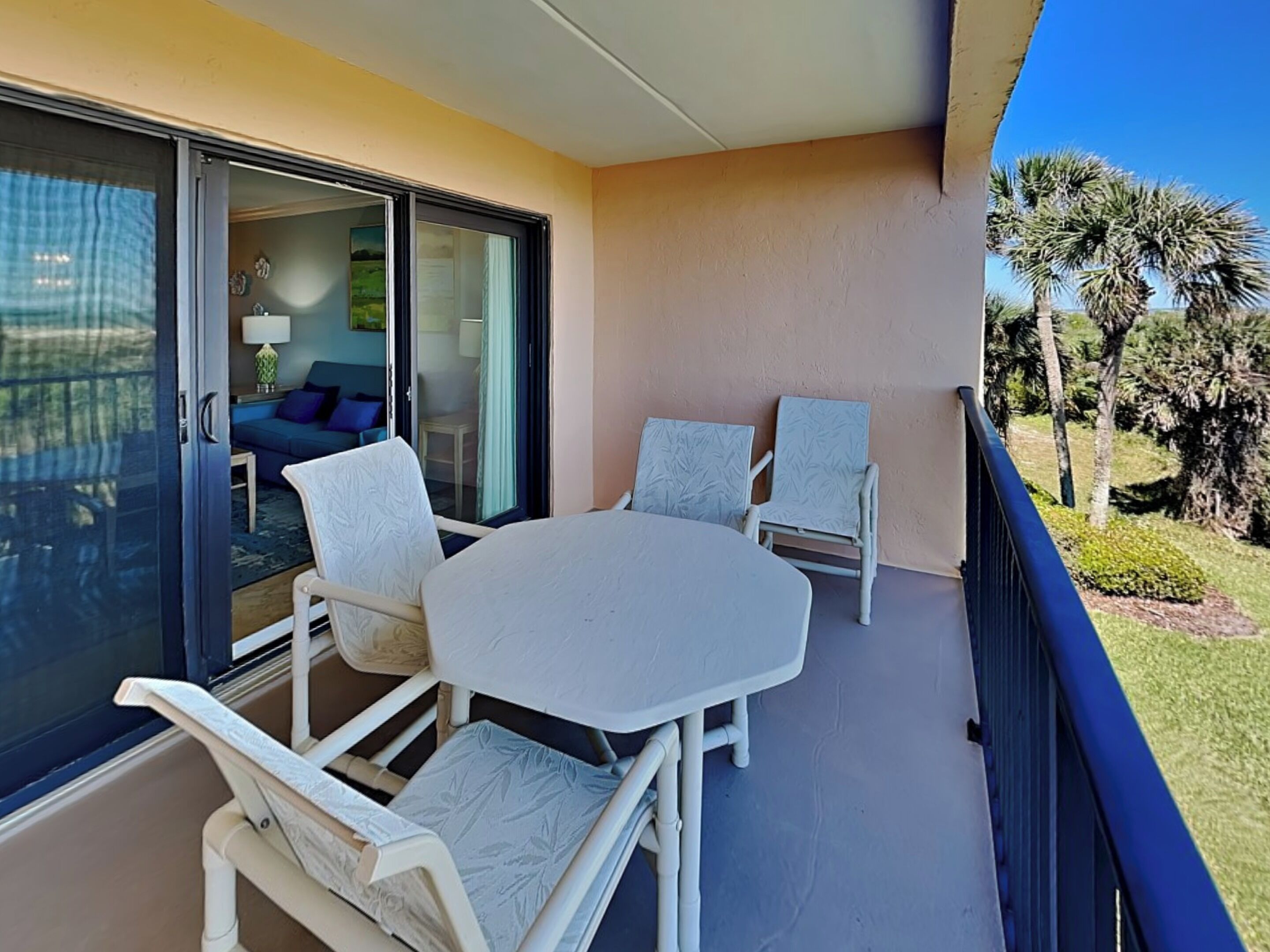 A balcony with a table and two chairs, a sliding glass door leading to a room with a sofa and a lamp, and a view of palm trees.