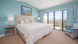 A bedroom with a large bed, a nightstand, a chair, and a view of the ocean through a balcony. - Sea Place by Distinctive Beach Rentals (St. Augustine)