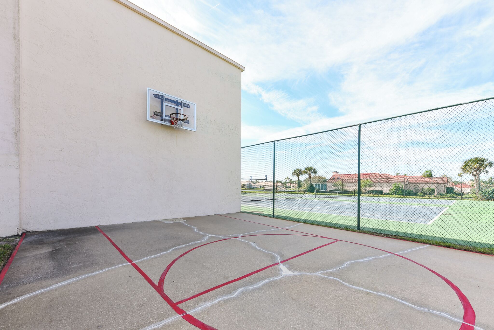 Una cancha de baloncesto al aire libre con una cerca de malla, aro de baloncesto y superficie de concreto con marcas rojas y blancas.