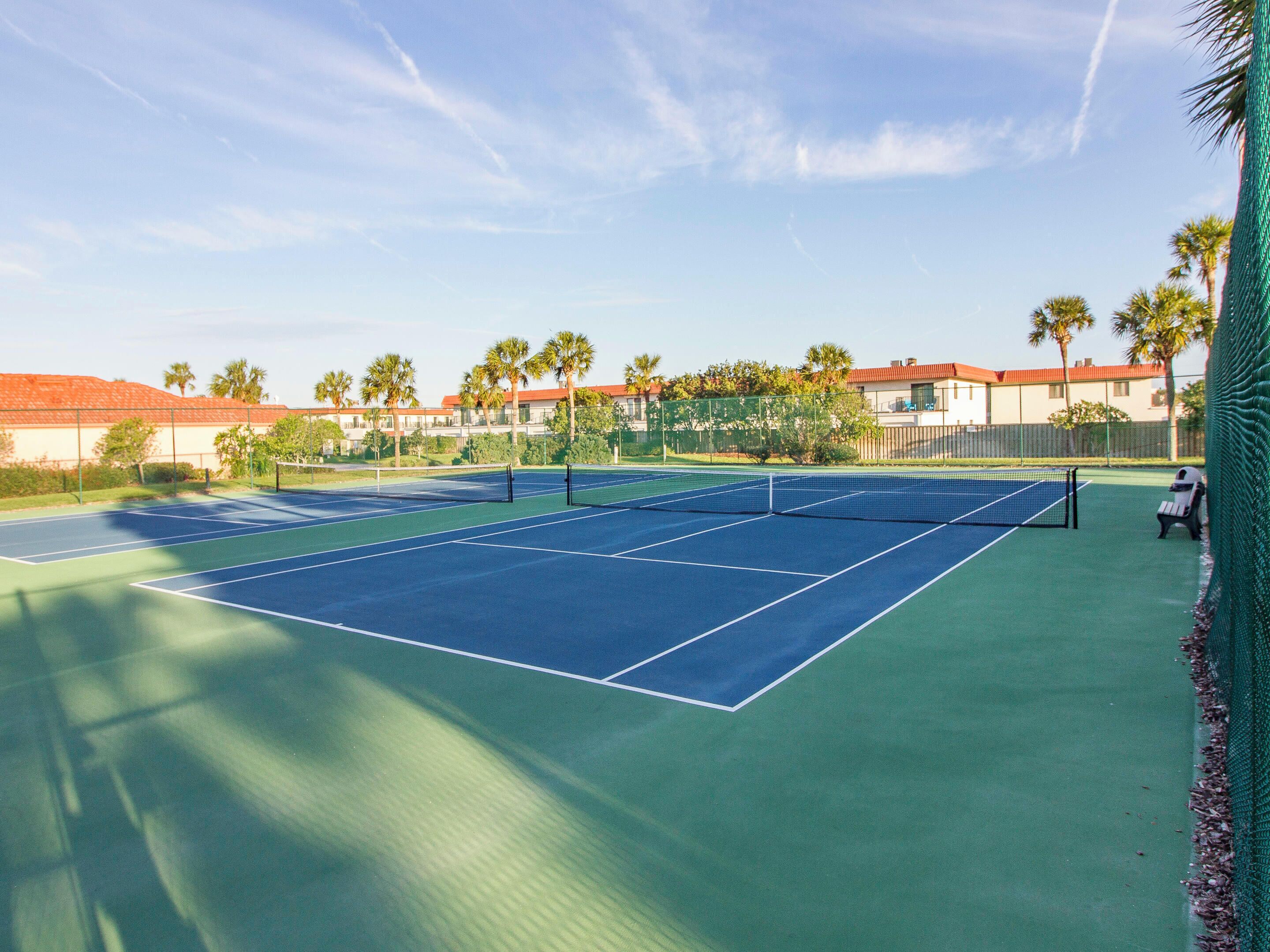 Un court de tennis avec un filet, entouré de palmiers et d’immeubles.