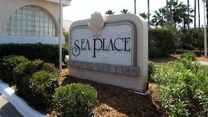A sign with the text "SEA PLACE" and a shell decoration, surrounded by greenery and palm trees. - Sea Place by Distinctive Beach Rentals (St. Augustine)