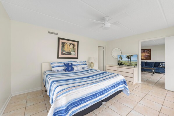 A bedroom with a bed, a ceiling fan, a painting, a dresser, a TV, and a view of a blue sofa and a wall with a circular mirror.