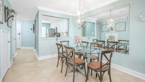 A dining area with a glass table surrounded by wooden chairs with woven seats, a modern chandelier, and a kitchen in the background. - Sea Place by Distinctive Beach Rentals (St. Augustine)