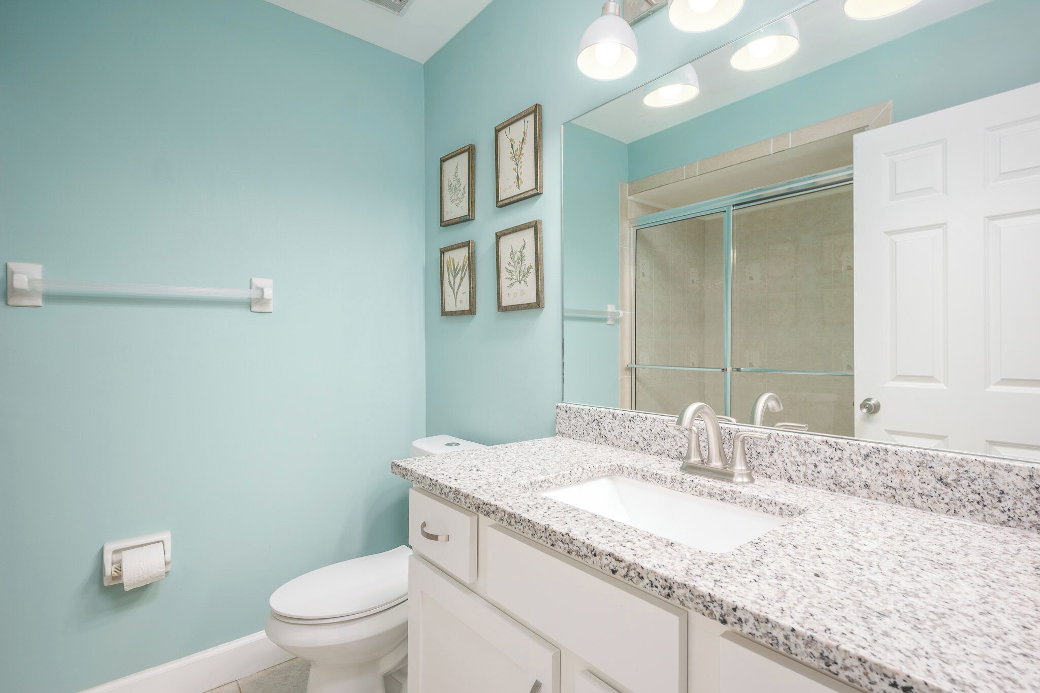 Une salle de bain avec des toilettes blanches, un lavabo sur un comptoir en granit, un meuble blanc, un porte-serviettes et des reproductions encadrées de motifs botaniques au mur.