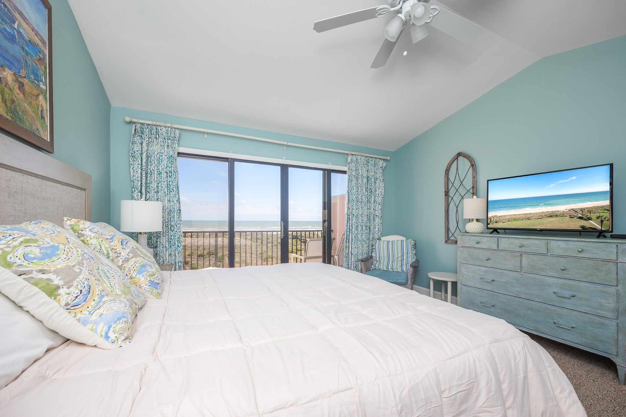 A bedroom with a large bed, a dresser, a television, and a view of the ocean through the window.