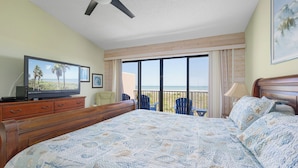 A hotel room with a large bed, a ceiling fan, a TV, a balcony with chairs, and a view of the beach. - Sea Place by Distinctive Beach Rentals (St. Augustine)