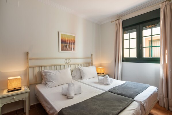 A double bed with white linens, a bedside table with a lamp, and a window with curtains.