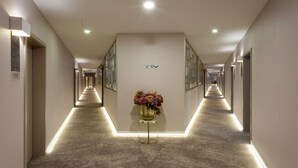 Un long couloir bien éclairé, agrémenté d’un vase décoratif au centre, de tableaux encadrés aux murs et d’une signalétique claire indiquant les directions.