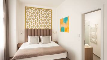 A modern hotel room with a large bed, a geometric wall panel, a small bedside table, and a bathroom visible through an open door.