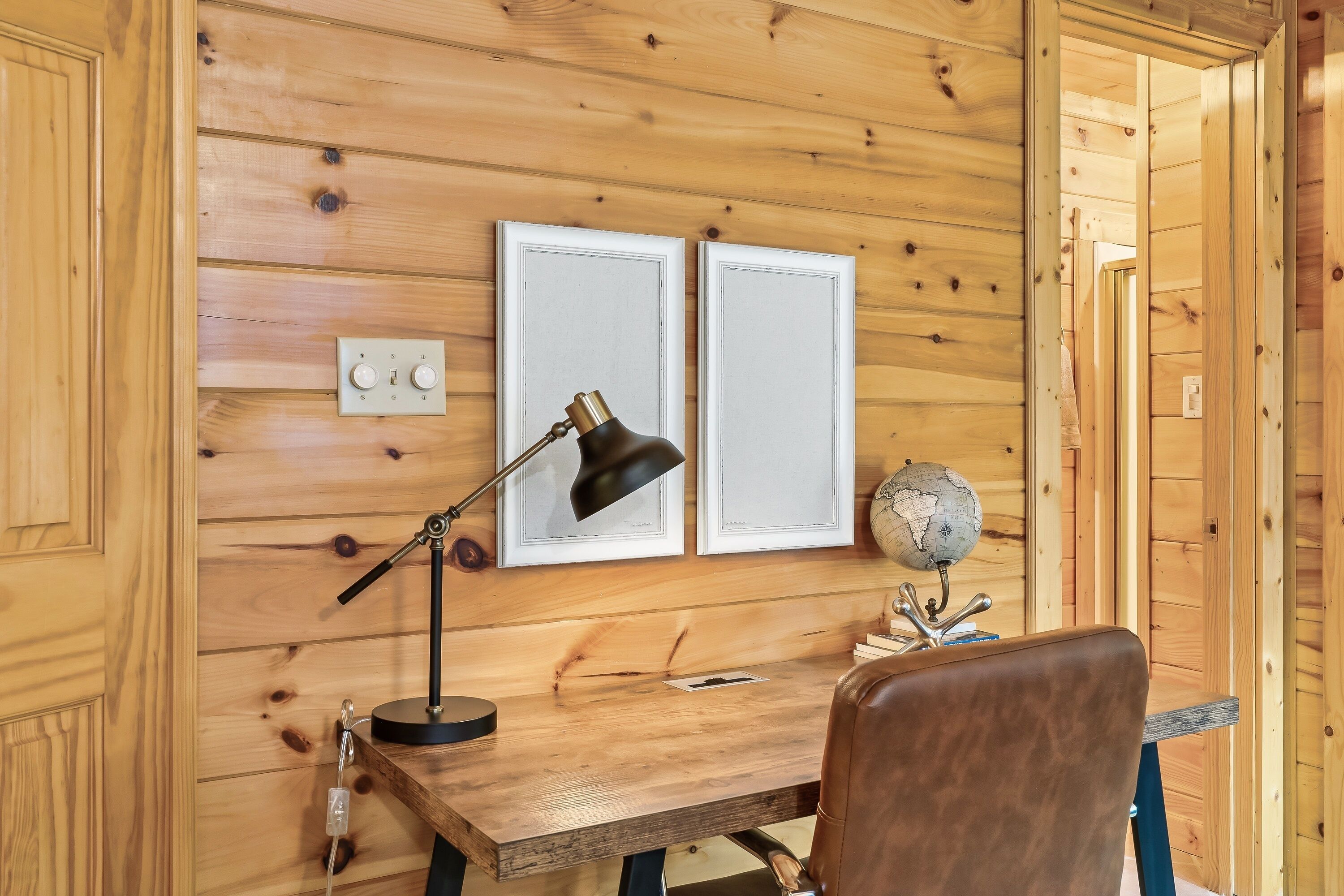 Une pièce aux murs lambrissés, comprenant un bureau, un fauteuil en cuir, un globe terrestre et deux tableaux encadrés.