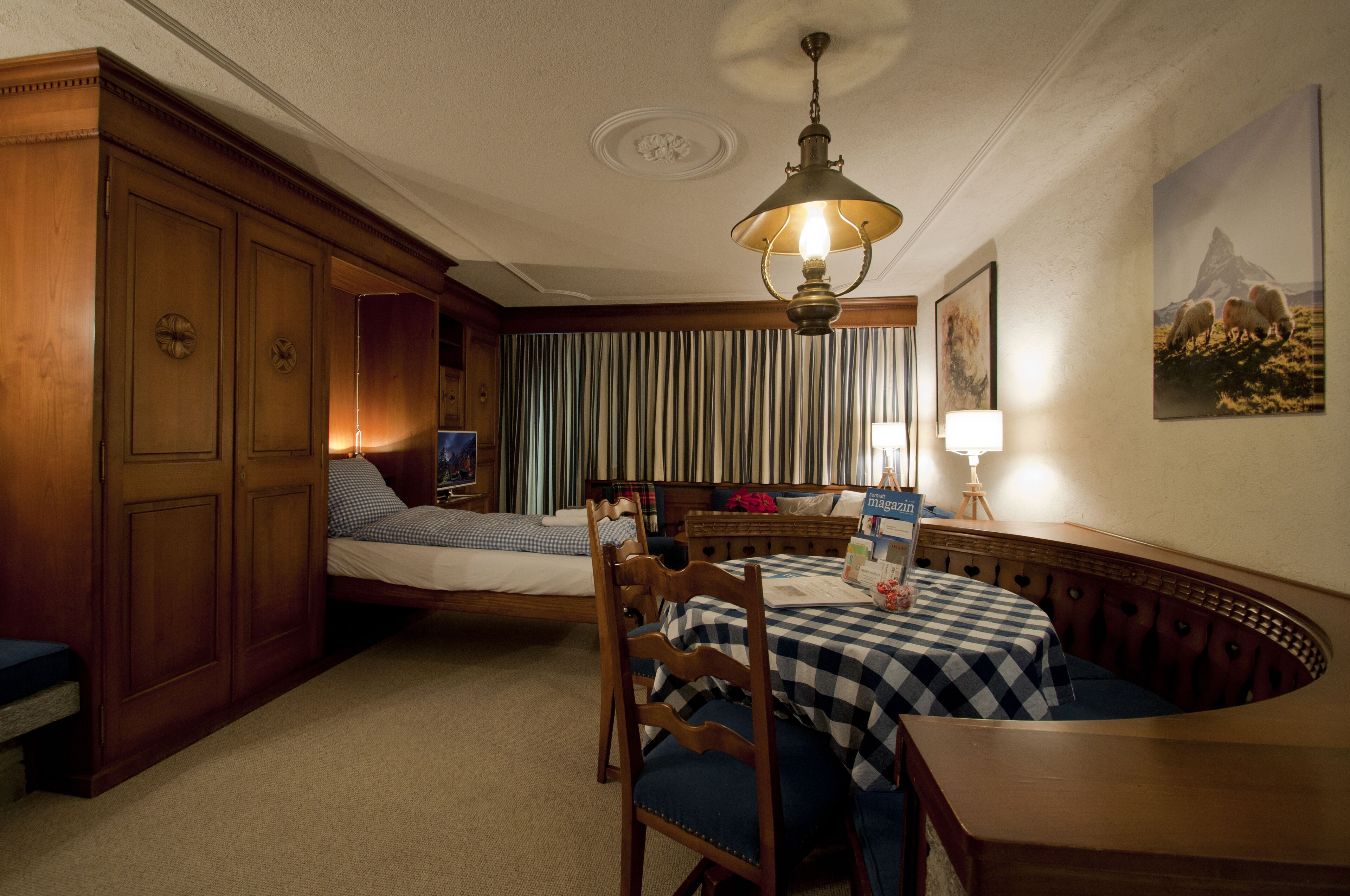 A hotel room with a bed, a desk with a chair, a dining table with a bench, and a large window with curtains.
