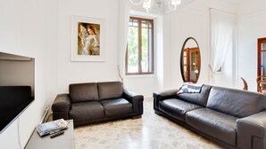 Ein Wohnzimmer mit zwei Ledersofas, einem Flachbildfernseher, einem Couchtisch mit Büchern und Fernbedienungen, einer Kristallleuchtre und einem großen Spiegel.