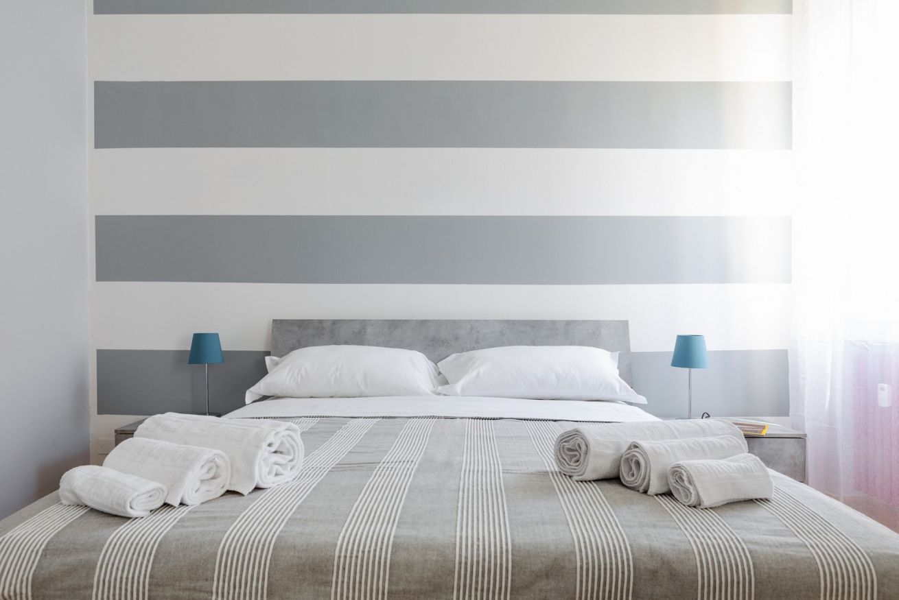 Een net opgemaakt bed met witte lakens en opgerolde handdoeken, een grijs gestreept beddegoed en een modern hoofdbord.