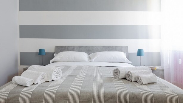 Una cama bien hecha con sábanas blancas y toallas enrolladas, una colcha a rayas grises y un cabecero moderno.