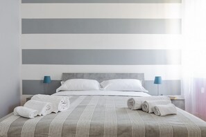 Uma cama bem arrumada com lençóis brancos e toalhas dobradas, um espalha cama cinza listrado e uma cabeceira moderna.