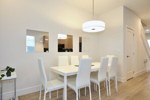 Un espace repas moderne avec une table et des chaises blanches, un luminaire suspendu et une vue sur la cuisine à travers une fenêtre.