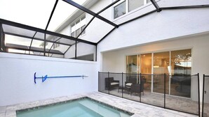 Une piscine entourée de parois en verre, un patio couvert aménagé avec du mobilier d’extérieur et une porte-fenêtre coulissante donnant sur un espace intérieur.